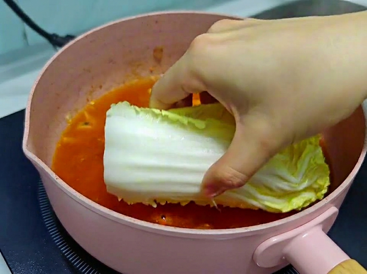 Une main déposant délicatement une grande feuille de chou chinois frais dans une poêle rose remplie de bouillon de kimchi frémissant.