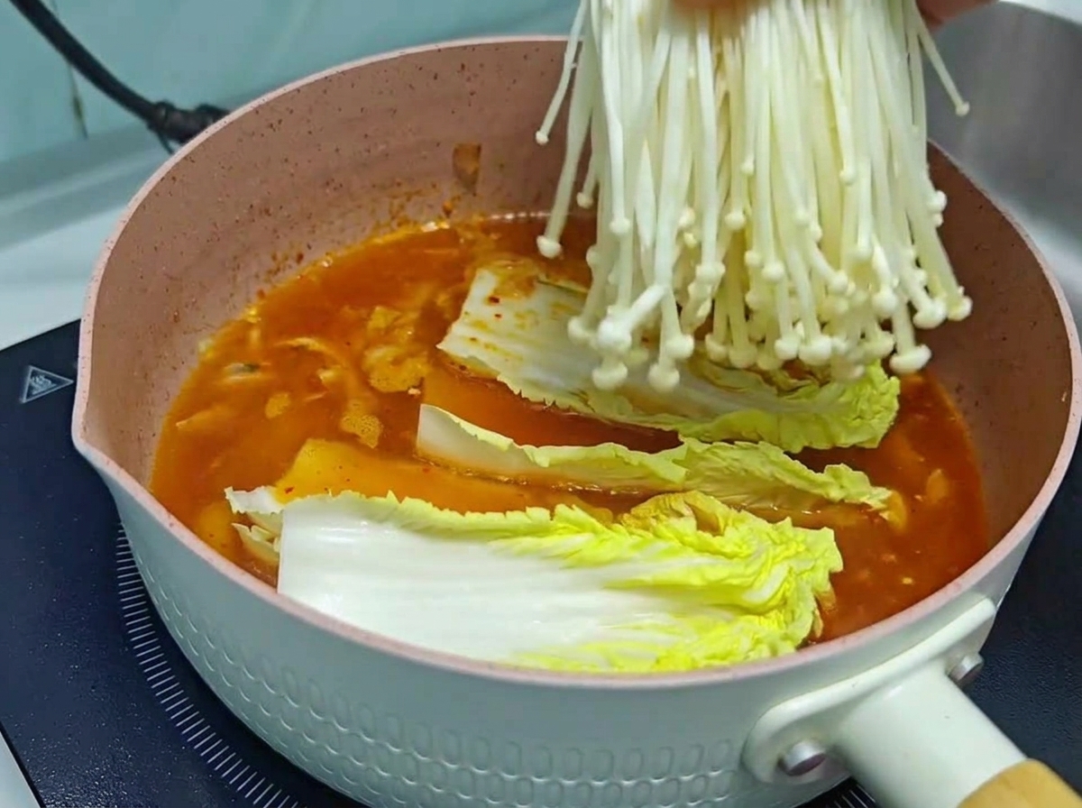 Gros plan dune main plongeant un bouquet frais de champignons enoki blancs dans une marmite rose contenant une base de soupe au kimchi rouge-orangé et du chou chinois.