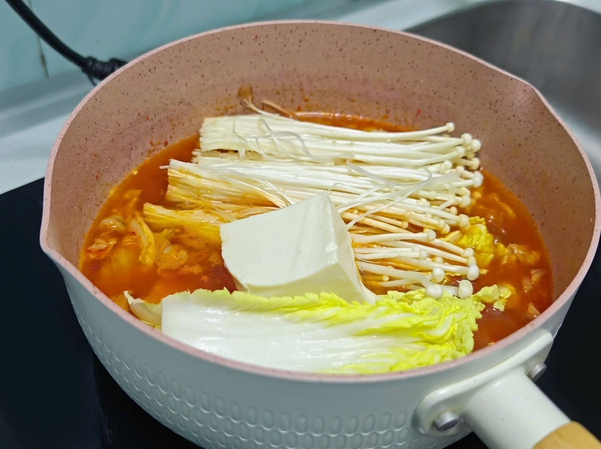 Un gros bloc de tofu blanc frais posé sur des champignons enoki et du chou dans une marmite de soupe au kimchi orange.