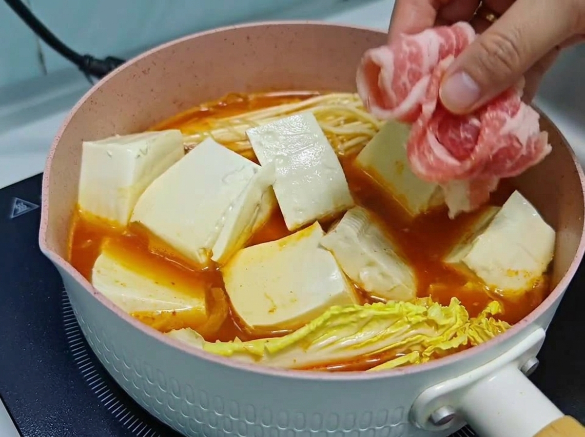 Une main superposant soigneusement de fins rouleaux de bœuf cru sur des cubes de tofu et des légumes dans une marmite de soupe au kimchi frémissante.