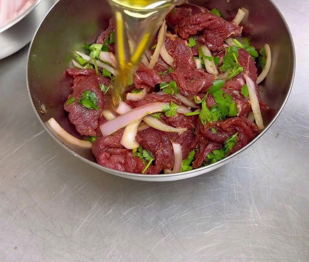 Une personne versant de lhuile de cuisson dans un bol en acier inoxydable rempli de bœuf assaisonné en cours de marinade, doignons émincés et de coriandre.