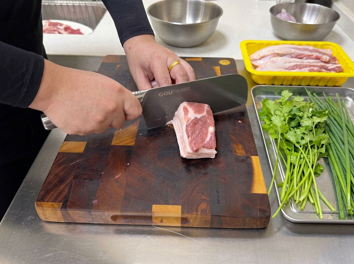 Une personne utilisant un grand couteau de cuisine pour couper un bloc de poitrine de porc crue et marbrée en fines lamelles sur une planche à découper.