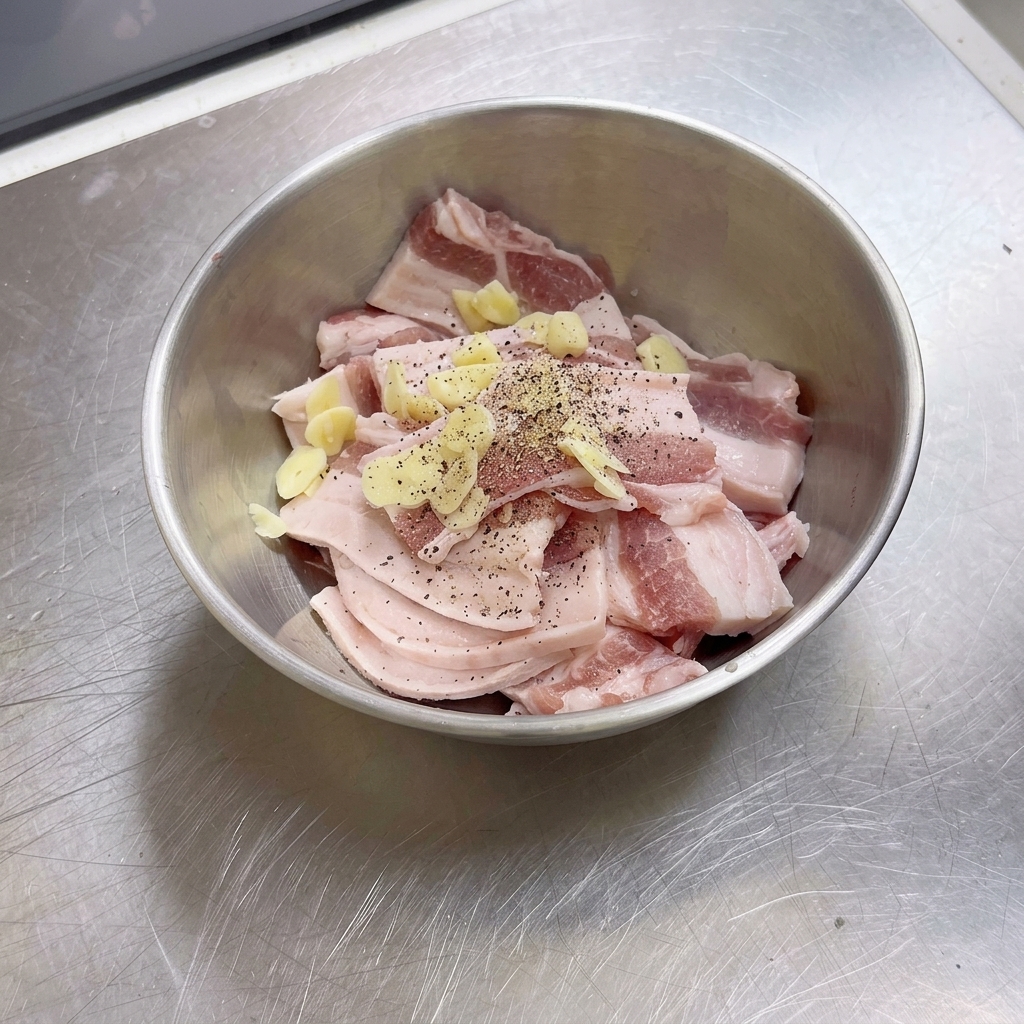 Un bol en acier inoxydable rempli de tranches de poitrine de porc crue, de morceaux dail et de poivre noir fraîchement moulu.