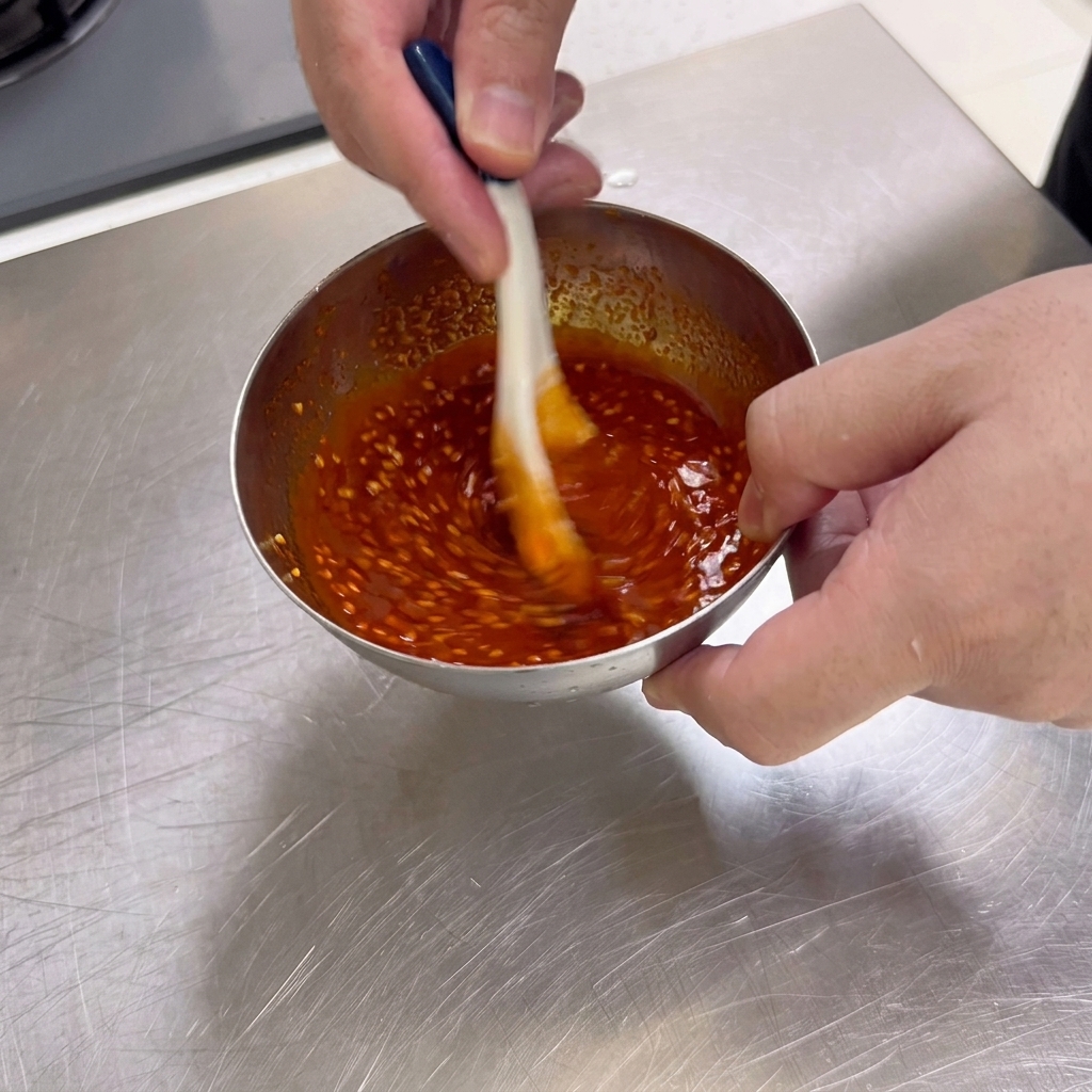Une sauce daccompagnement pour barbecue, épaisse et rouge foncé, parsemée de graines de sésame, remuée avec une cuillère dans un petit bol en acier inoxydable.