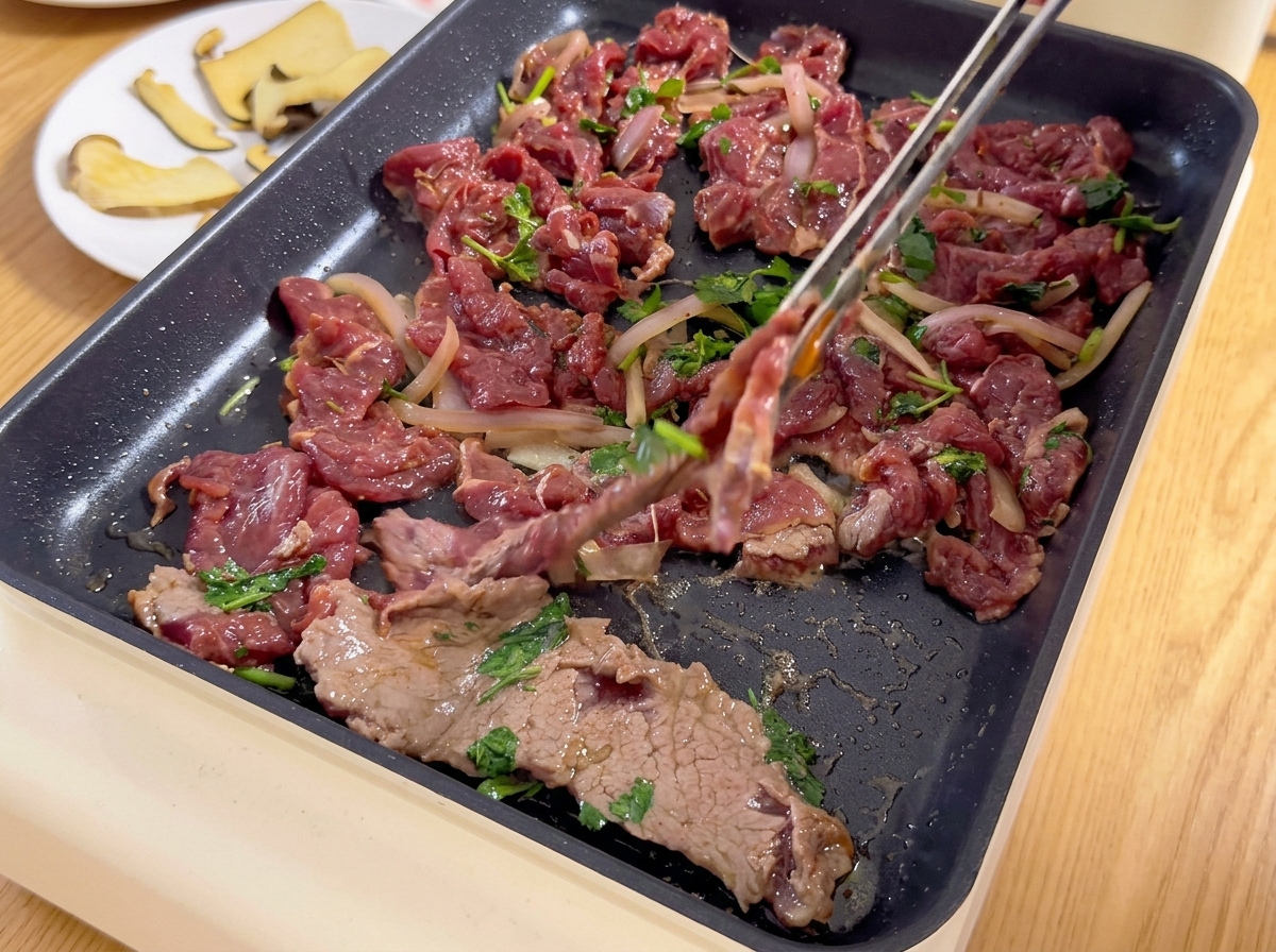 De fines tranches de bœuf mariné mélangées à des oignons et de la coriandre déposées sur une plancha électrique chaude à laide dune pince.