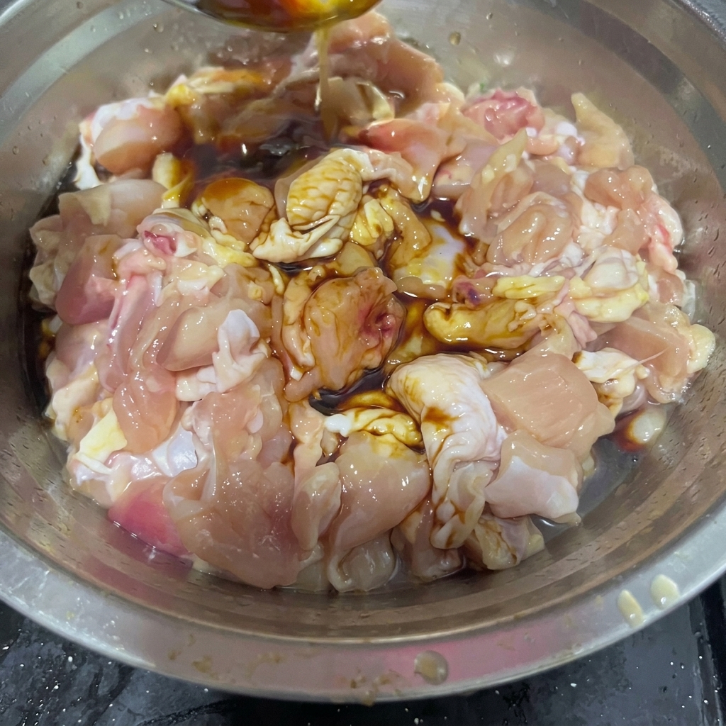Vin de cuisine versé depuis une cuillère en acier inoxydable dans un bol contenant des morceaux de poulet cru et des sauces sombres.