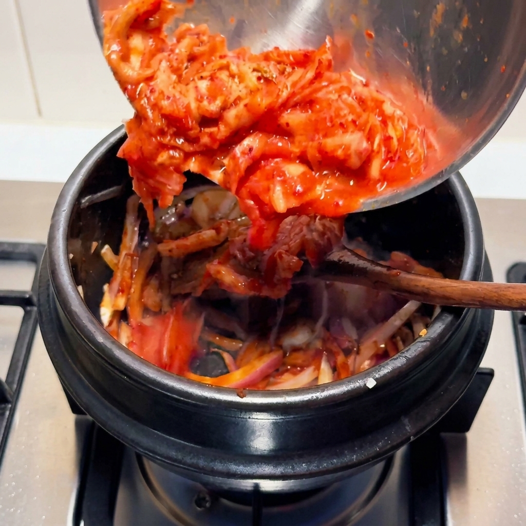Kimchi fermenté rouge vif et haché versé depuis un bol en acier inoxydable dans un pot de cuisson en pierre noire sur une cuisinière.