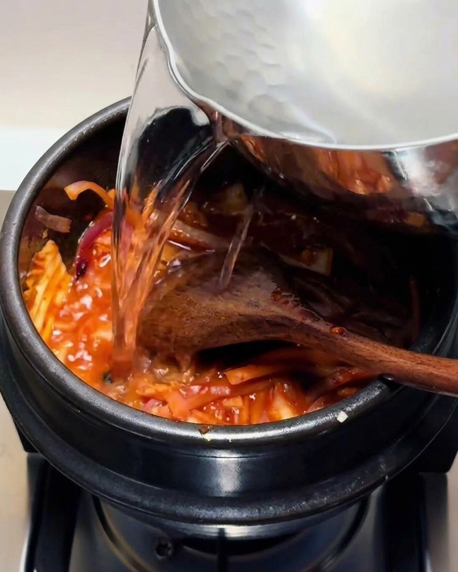 De leau chaude claire versée depuis une casserole en métal dans un pot en pierre noire contenant le mélange de kimchi rouge.