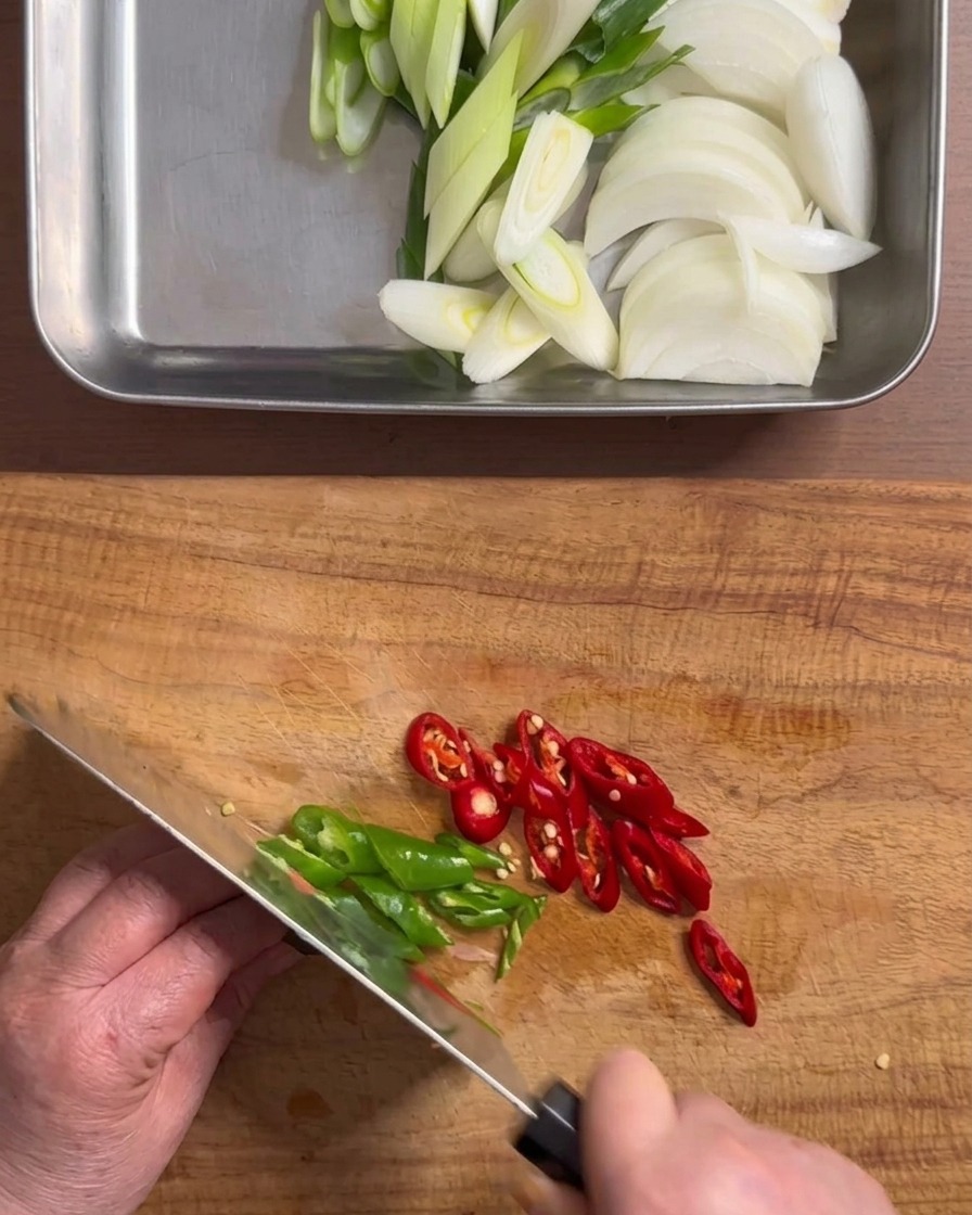 Mains coupant des piments verts et rouges en fines rondelles sur une planche à découper en bois.