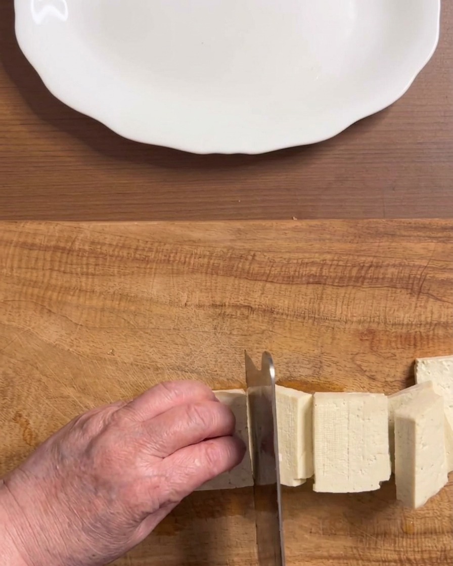 Mains coupant un bloc de tofu blanc ferme en morceaux rectangulaires réguliers sur une planche en bois.