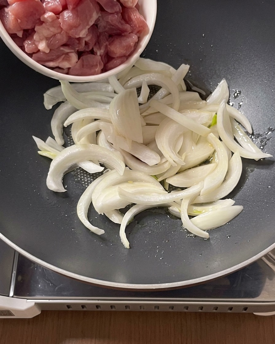 Morceaux de porc cru ajoutés depuis un bol dans une poêle contenant les oignons émincés et lhuile.