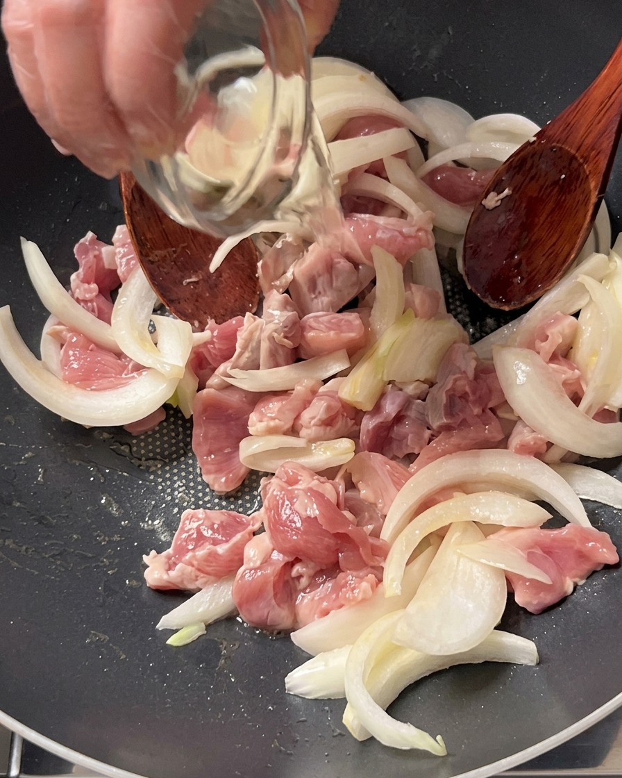 Vin de cuisson versé depuis un petit bol en verre sur le porc et les oignons en train de cuire.