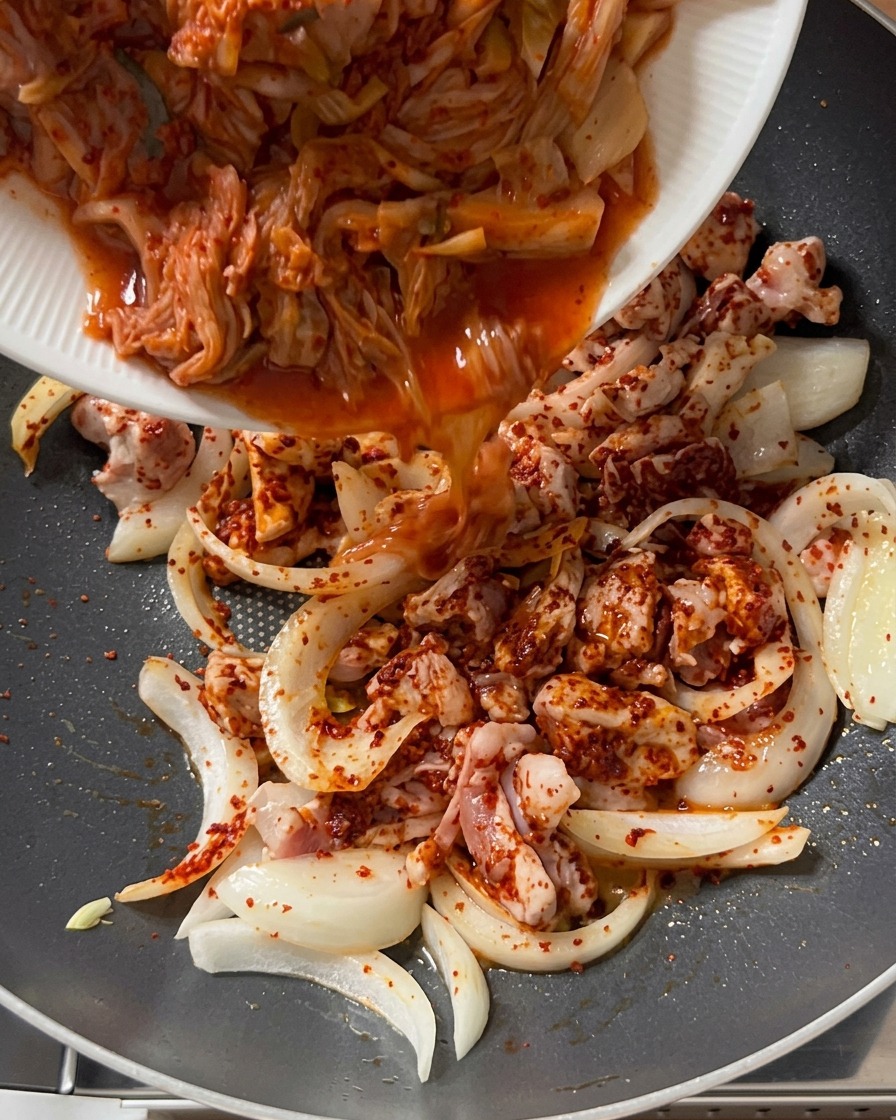 Kimchi coupé versé depuis une assiette blanche dans la poêle contenant le porc et les oignons bien assaisonnés.