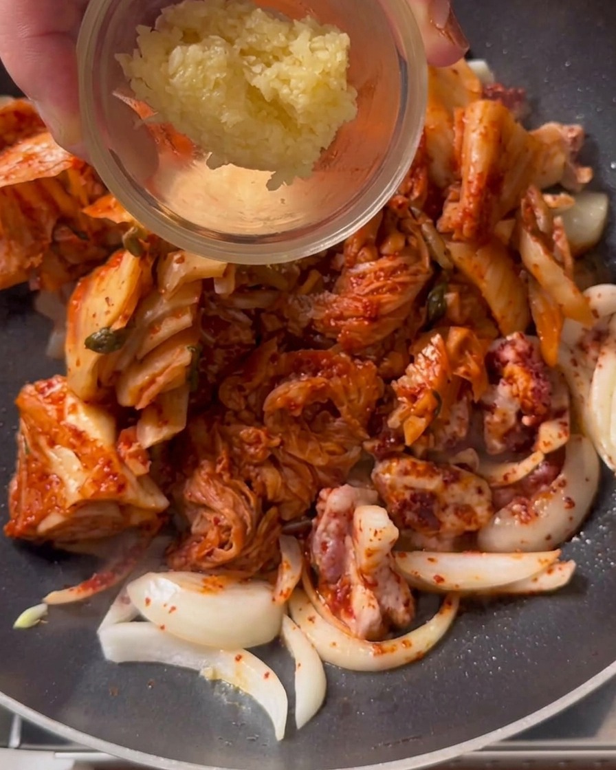 Une cuillère dail émincé ajoutée dans la poêle au milieu du kimchi et du porc sautés.