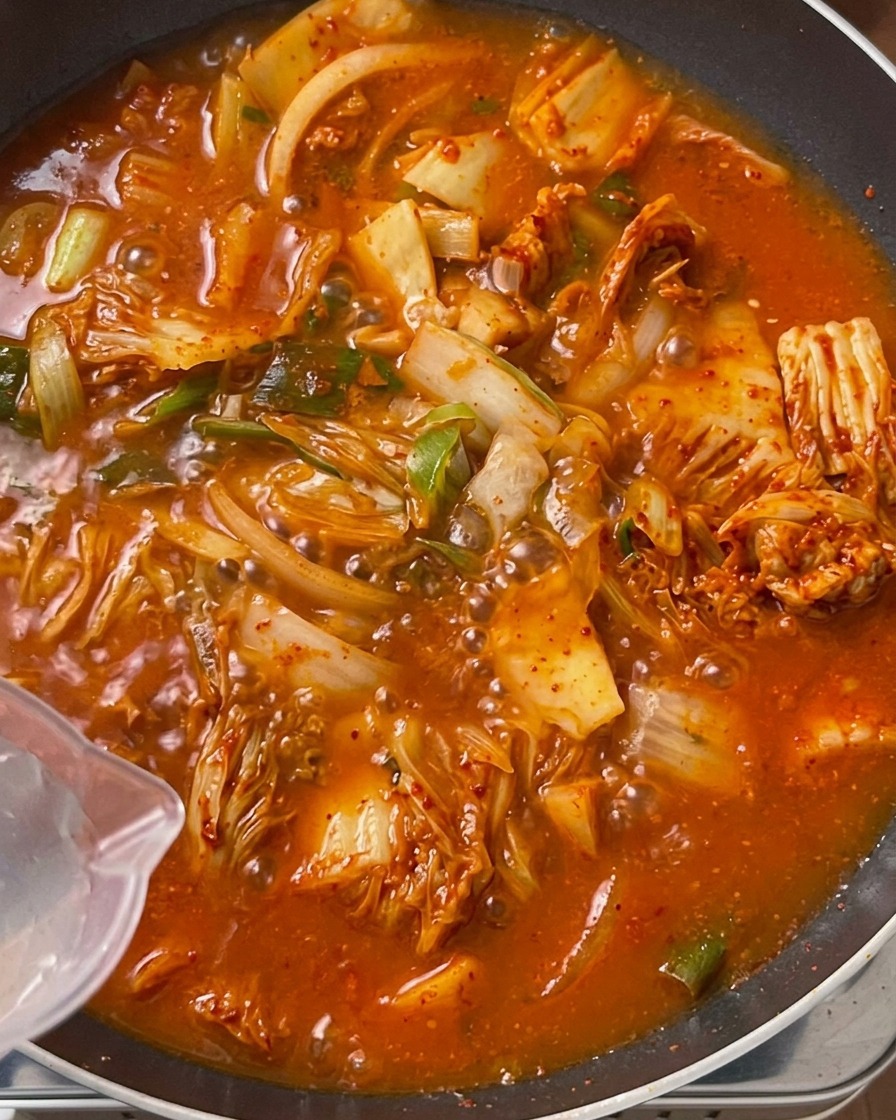 Eau claire versée depuis une carafe en plastique dans une grande sauteuse remplie dun mélange frémissant de kimchi et de porc dun rouge orangé éclatant.
