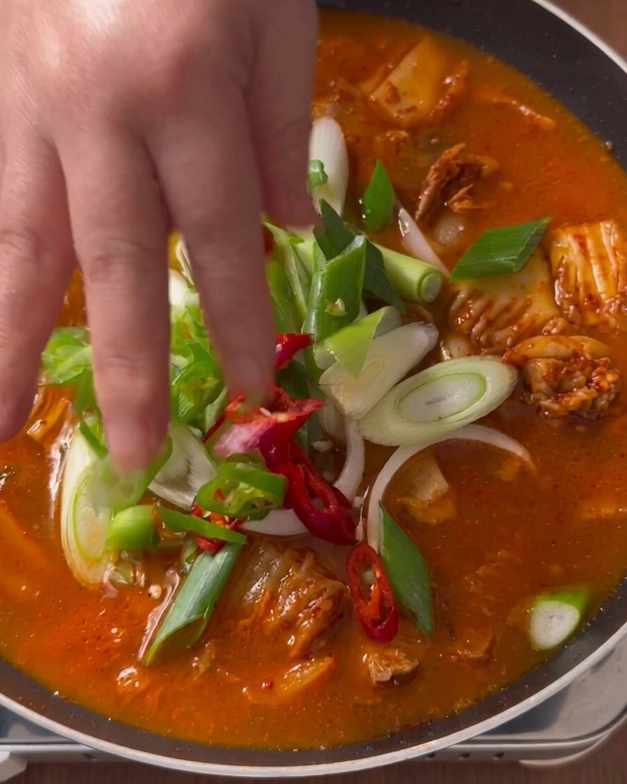 Une main déposant des oignons verts émincés et des rondelles de piments rouges et verts sur le dessus dun ragoût de kimchi rouge qui mijote.