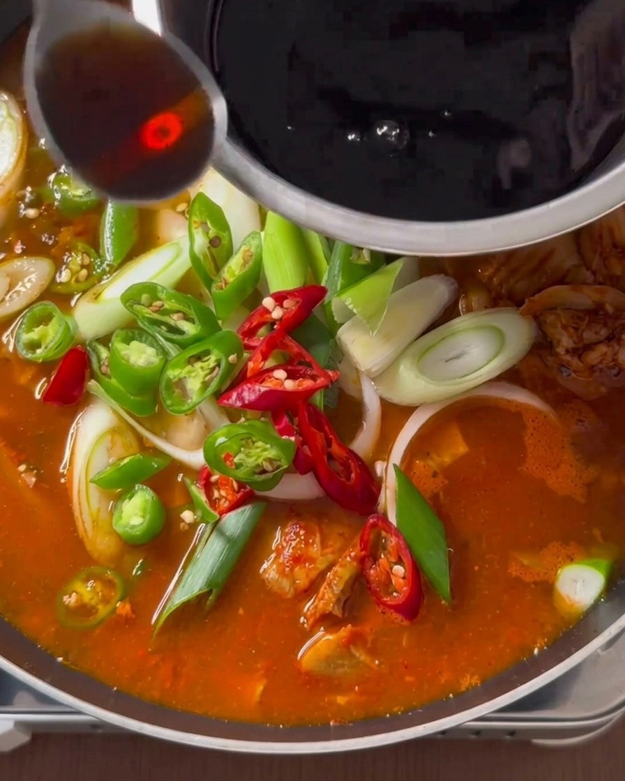 Une cuillère de sauce sombre versée depuis un bol en inox sur un ragoût de kimchi frémissant garni de piments et doignons verts.