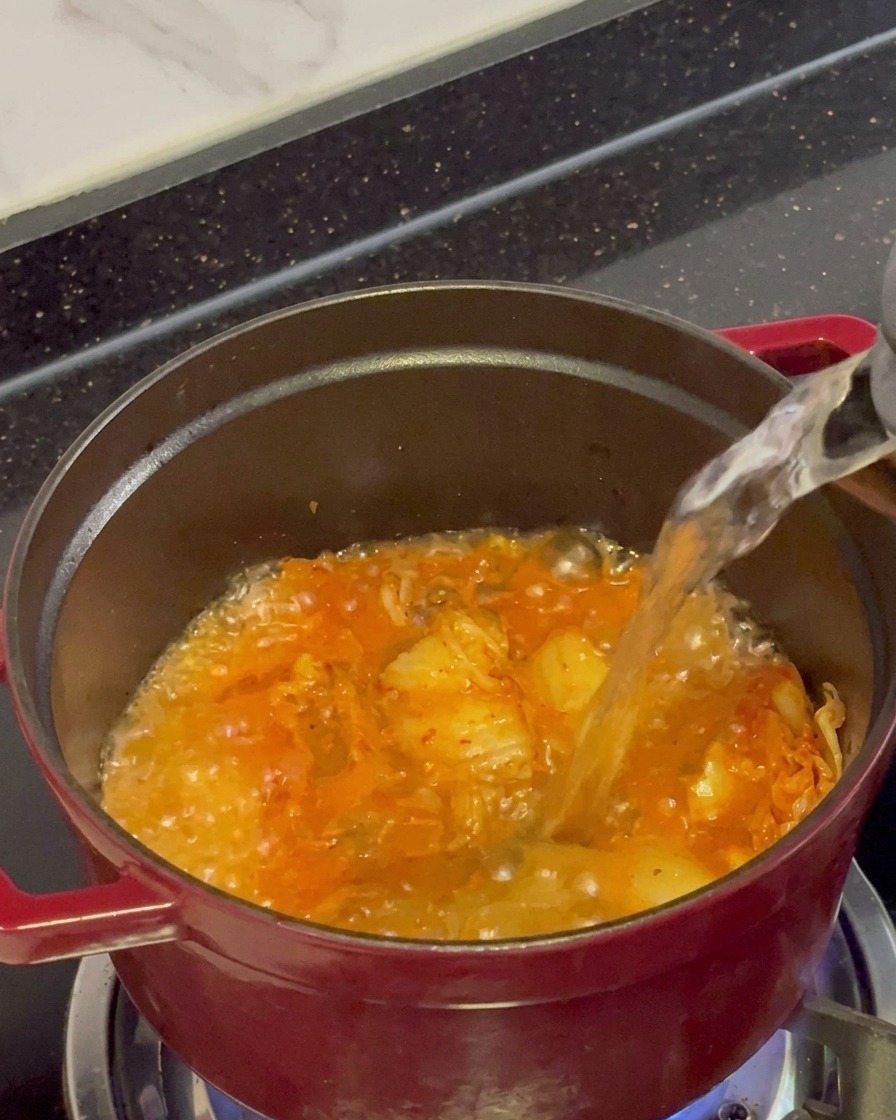 Eau bouillante claire versée dune bouilloire dans une marmite rouge remplie de kimchi frémissant et sauté.