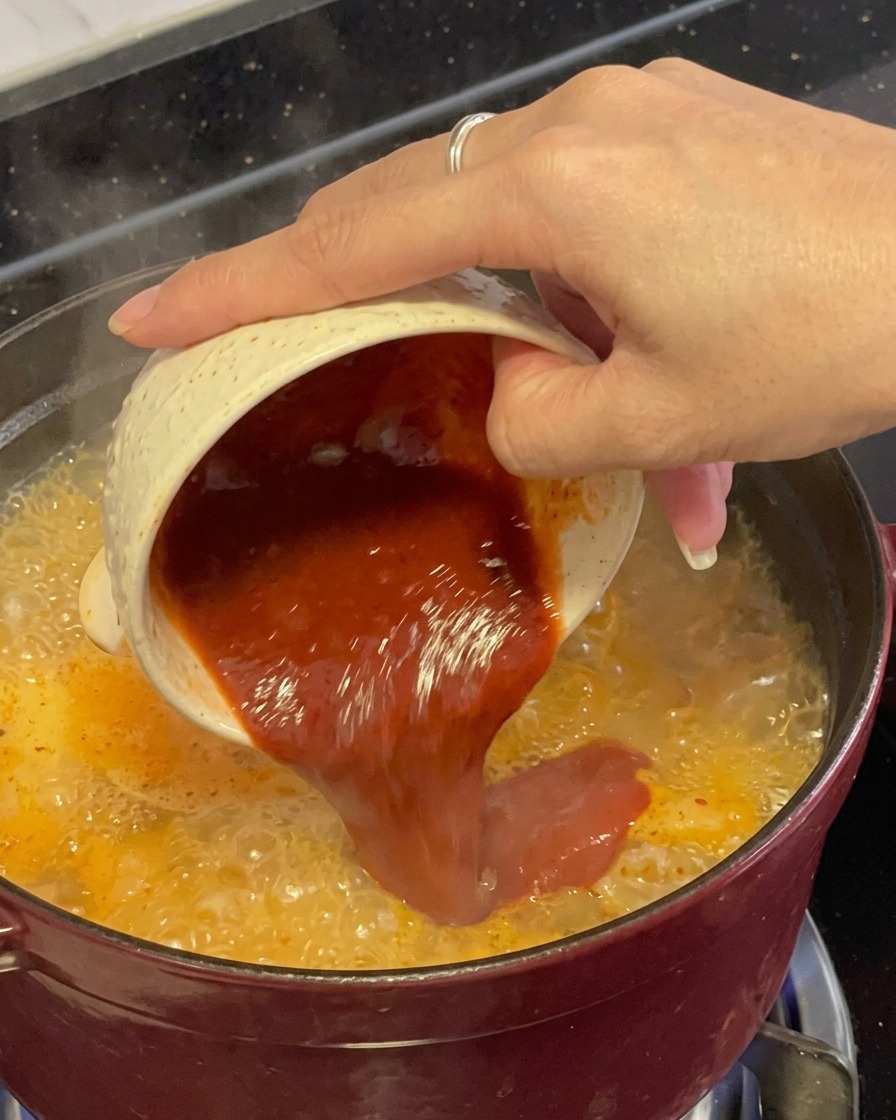 Mélange dassaisonnement au piment rouge foncé et épais versé depuis un petit bol moucheté directement dans un ragoût de fruits de mer rouge bouillonnant.