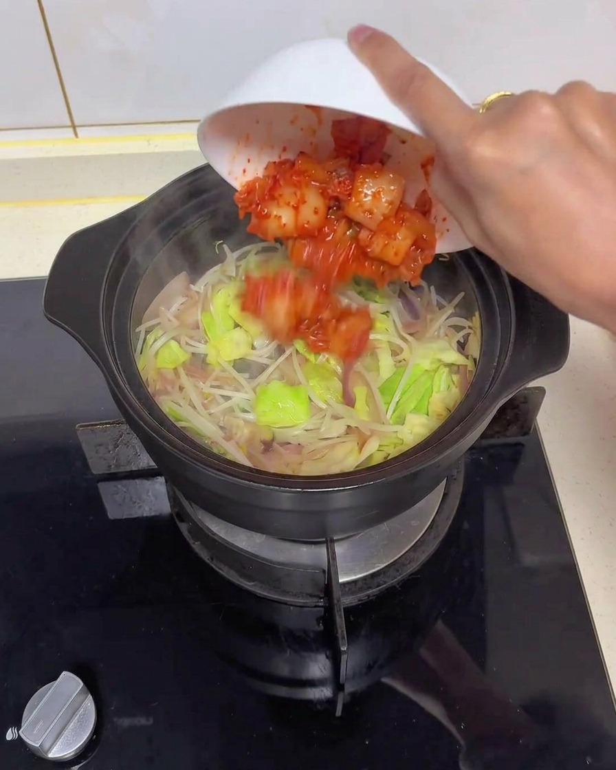 Une main penchant un bol blanc pour ajouter du kimchi rouge émincé dans une marmite en céramique noire remplie doignons translucides, de chou vert et de pousses de haricots.