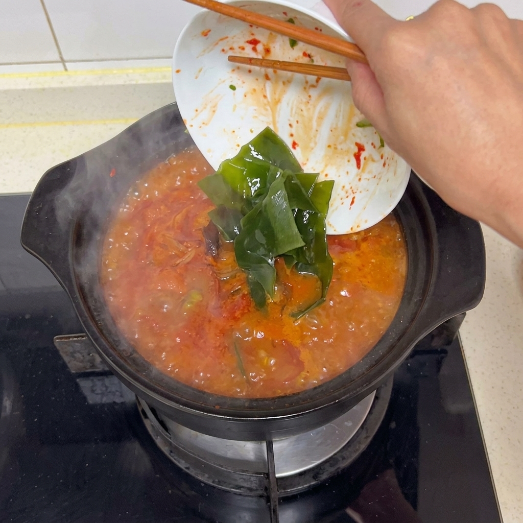 Des baguettes glissant de larges lanières de varech vert foncé réhydraté depuis un bol blanc vers un bouillon rouge vif frémissant.