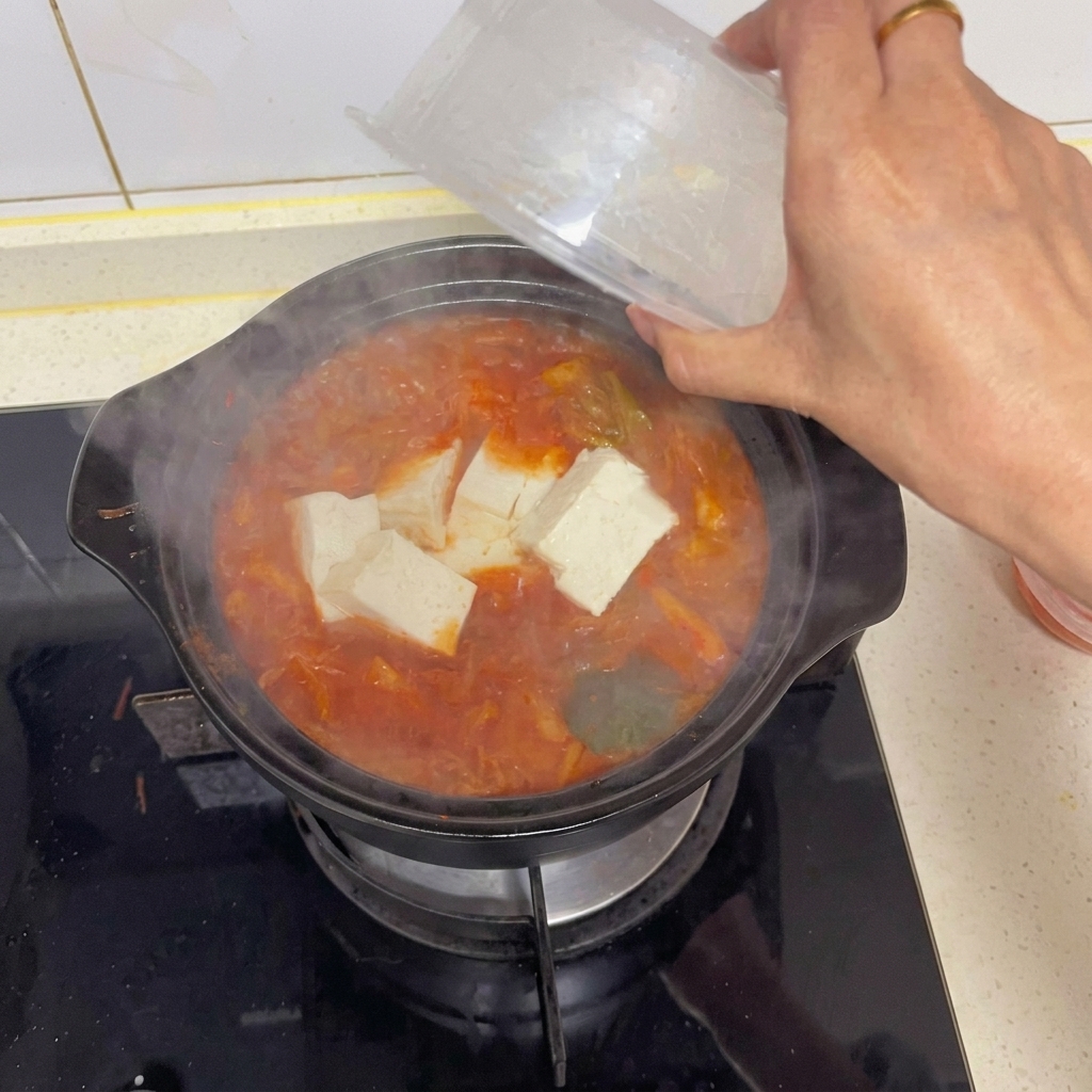 Une main ajoutant délicatement de grands cubes de tofu blanc depuis un récipient en plastique transparent dans une marmite de soupe au kimchi bouillonnante.