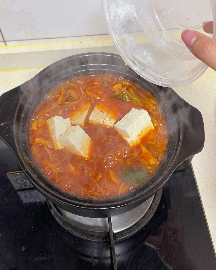 Blocs de tofu blanc rectangulaires plongés depuis un récipient transparent dans une marmite bouillonnante de soupe au kimchi rouge foncé contenant des légumes.