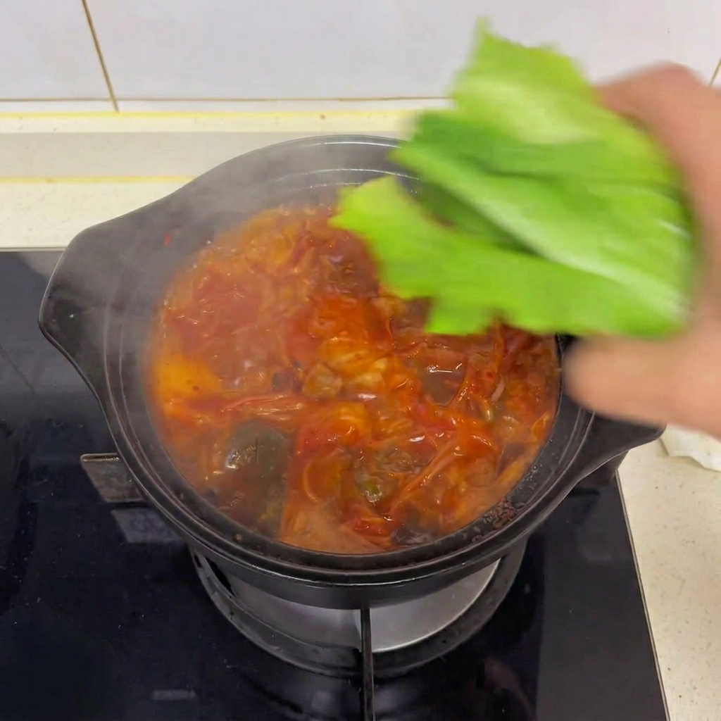 Une main déposant des feuilles de laitue romaine fraîche dans une marmite fumante de soupe au kimchi et tofu.