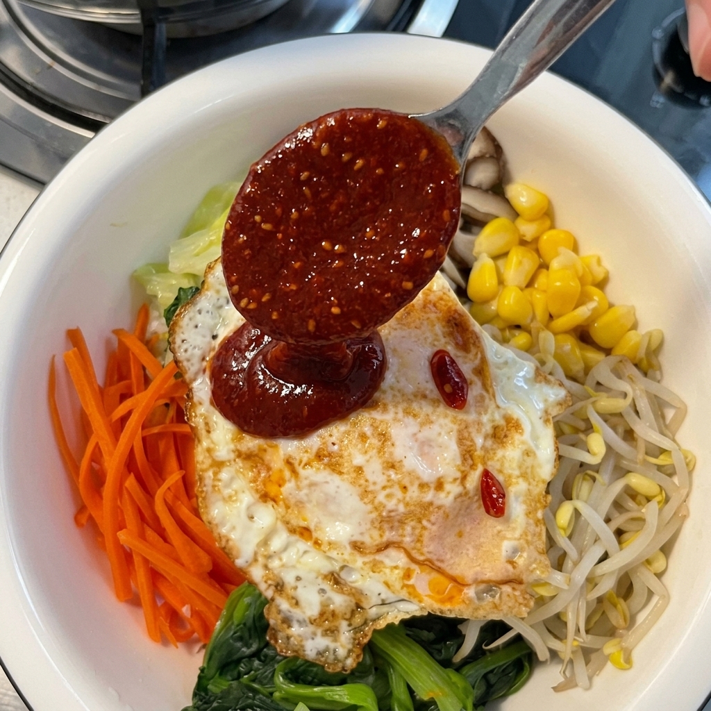 Une cuillère en métal versant une sauce épaisse à la pâte de piment rouge sur un œuf au plat disposé sur un assortiment coloré de légumes pour bibimbap.