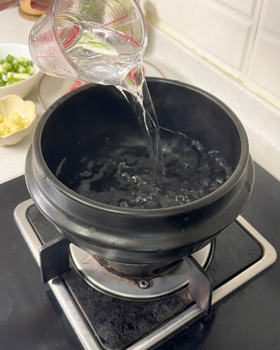 De leau versée depuis un verre doseur transparent dans une marmite en terre cuite noire sur un brûleur de cuisinière à gaz.