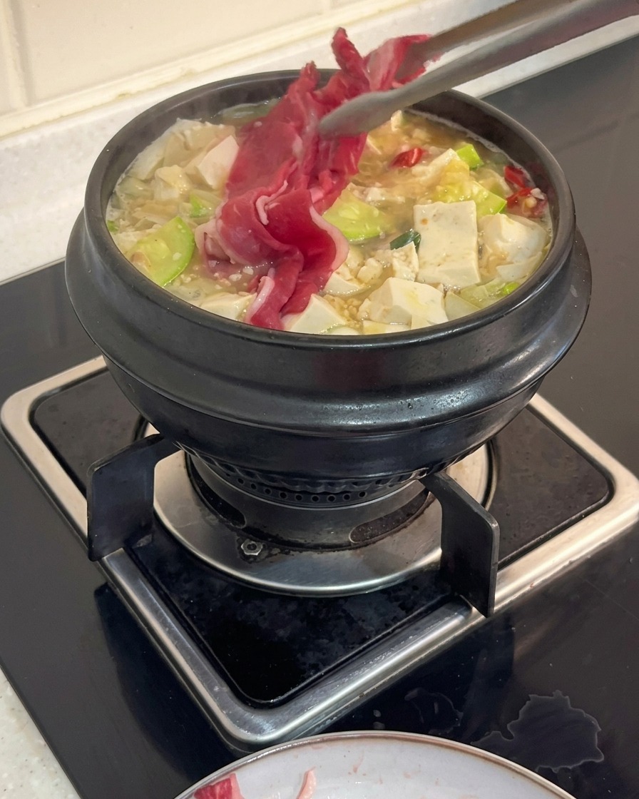 Une pince de cuisine déposant de fines tranches de poitrine de bœuf crue dans une marmite de ragoût coréen bouillant garni de tofu et de courgettes.