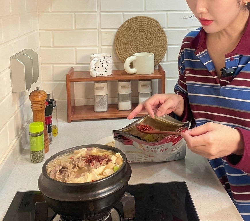 Une cuillérée de piment rouge en poudre grossier ajoutée dans une marmite en terre cuite bouillonnante de ragoût coréen contenant du tofu et du bœuf émincé.