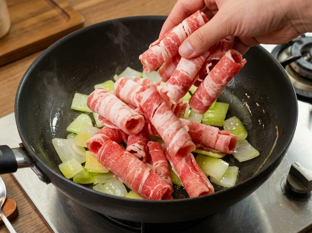 Fines tranches de bœuf cru ajoutées dans une poêle contenant des oignons revenus translucides.