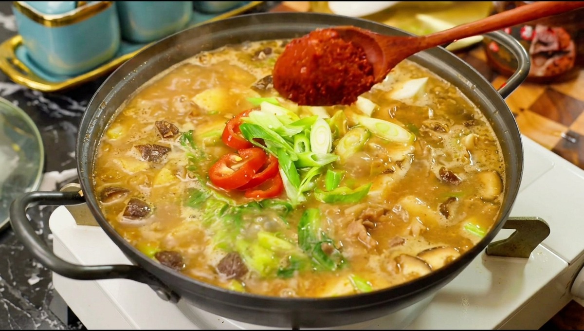 Une cuillère en bois versant une bonne quantité de poudre de piment rouge vif dans une marmite bouillonnante de ragoût de soja coréen avec du bœuf, des champignons et des piments frais.