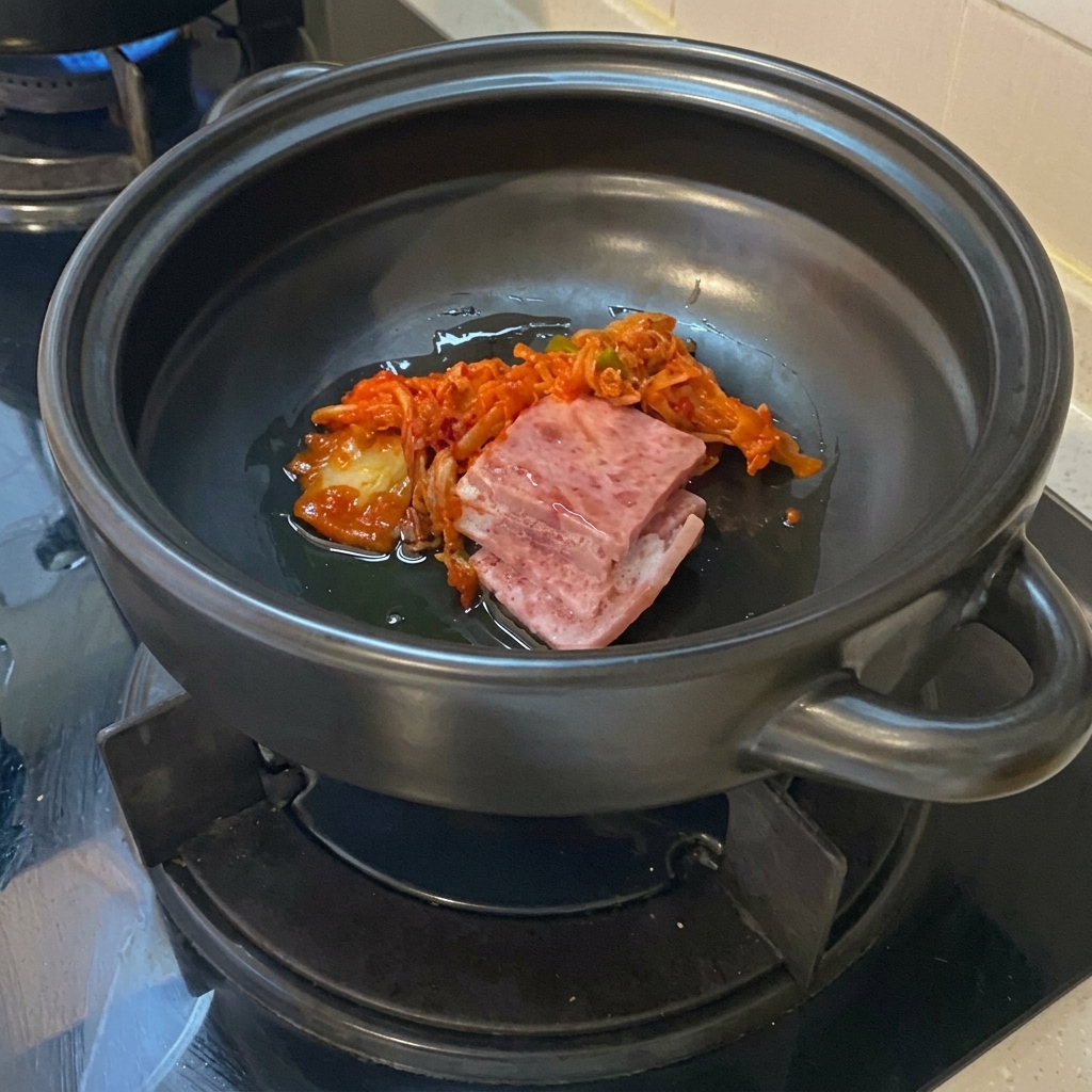 Tranches de poitrine de porc crue et kimchi haché coloré reposant dans lhuile au fond dune marmite en céramique noire.