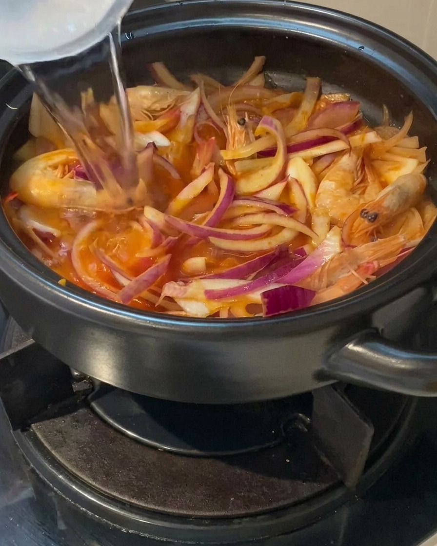 Un filet deau claire versé depuis une tasse dans une marmite en terre cuite remplie doignons rouges émincés et de crevettes entières.