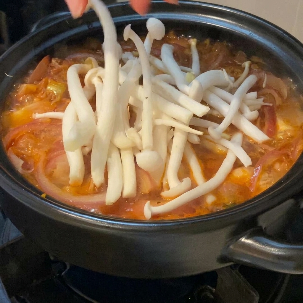 Champignons blancs frais délicatement déposés à la main sur une soupe au kimchi rouge et bouillonnante dans une marmite en terre cuite noire.