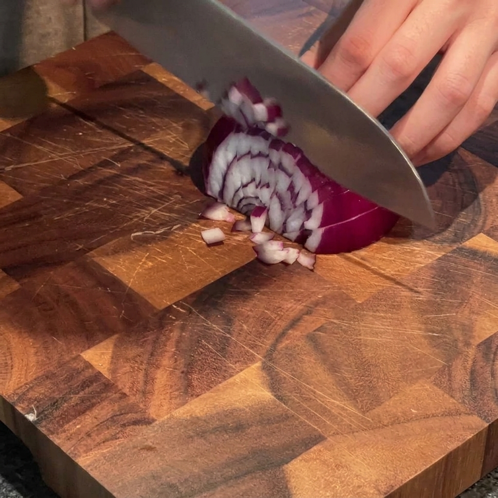 Une main utilisant un couteau de chef pour couper un oignon rouge en petits dés sur une planche en bois.