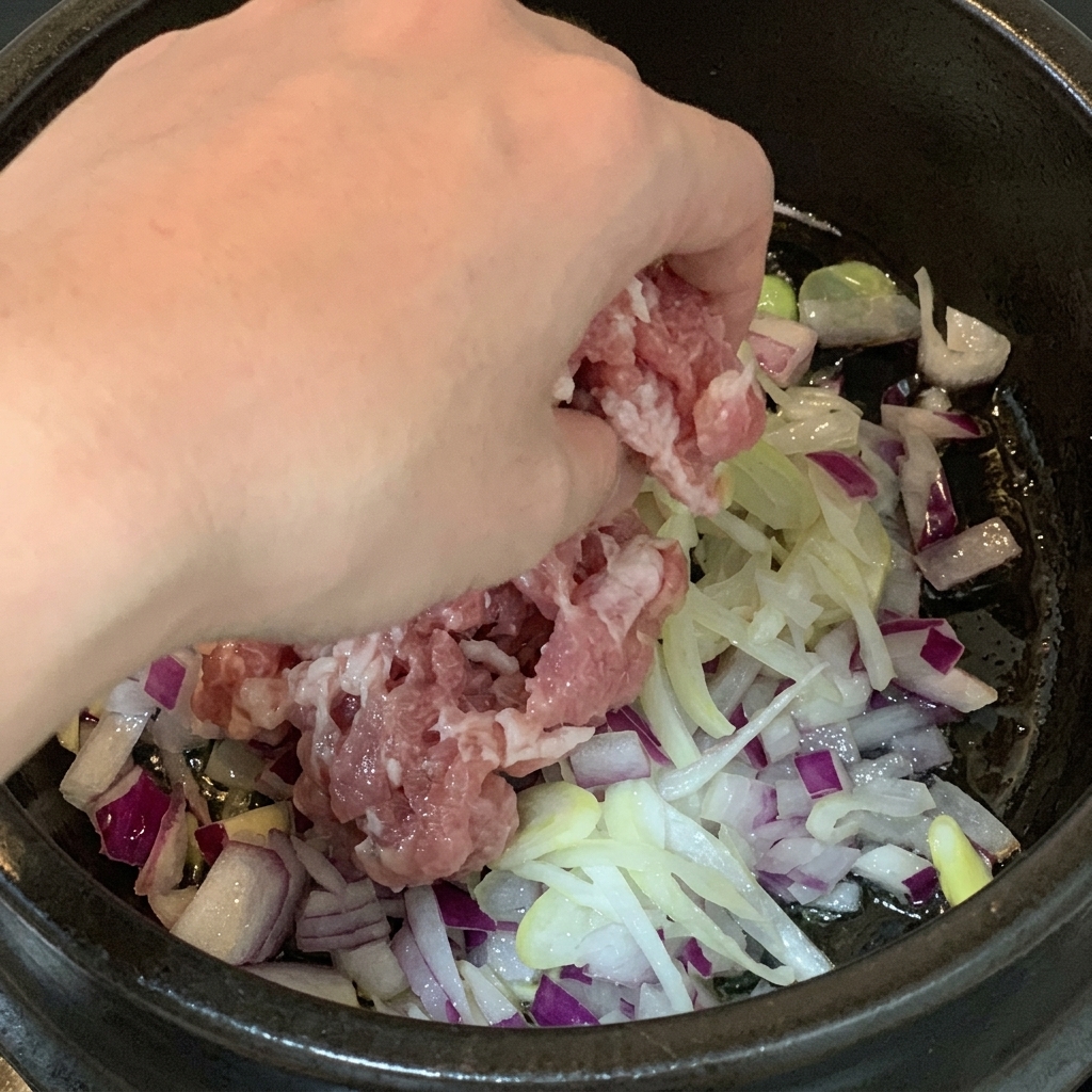 Morceaux de poitrine de porc crue ajoutés dans une marmite contenant des dés doignon rouge et des oignons verts émincés.