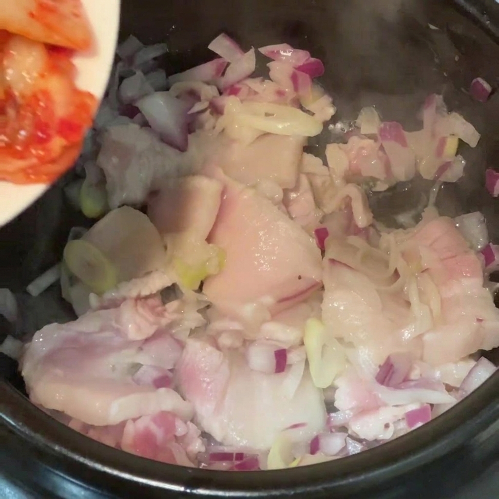 Chou kimchi coupé versé depuis une assiette dans une marmite contenant des morceaux de poitrine de porc et des oignons.