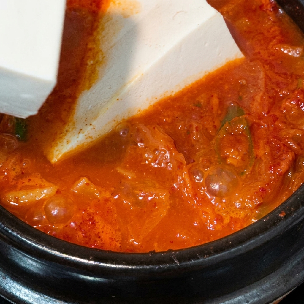 Un grand bloc de tofu soyeux blanc délicatement déposé dans une marmite de soupe de kimchi rouge en ébullition.