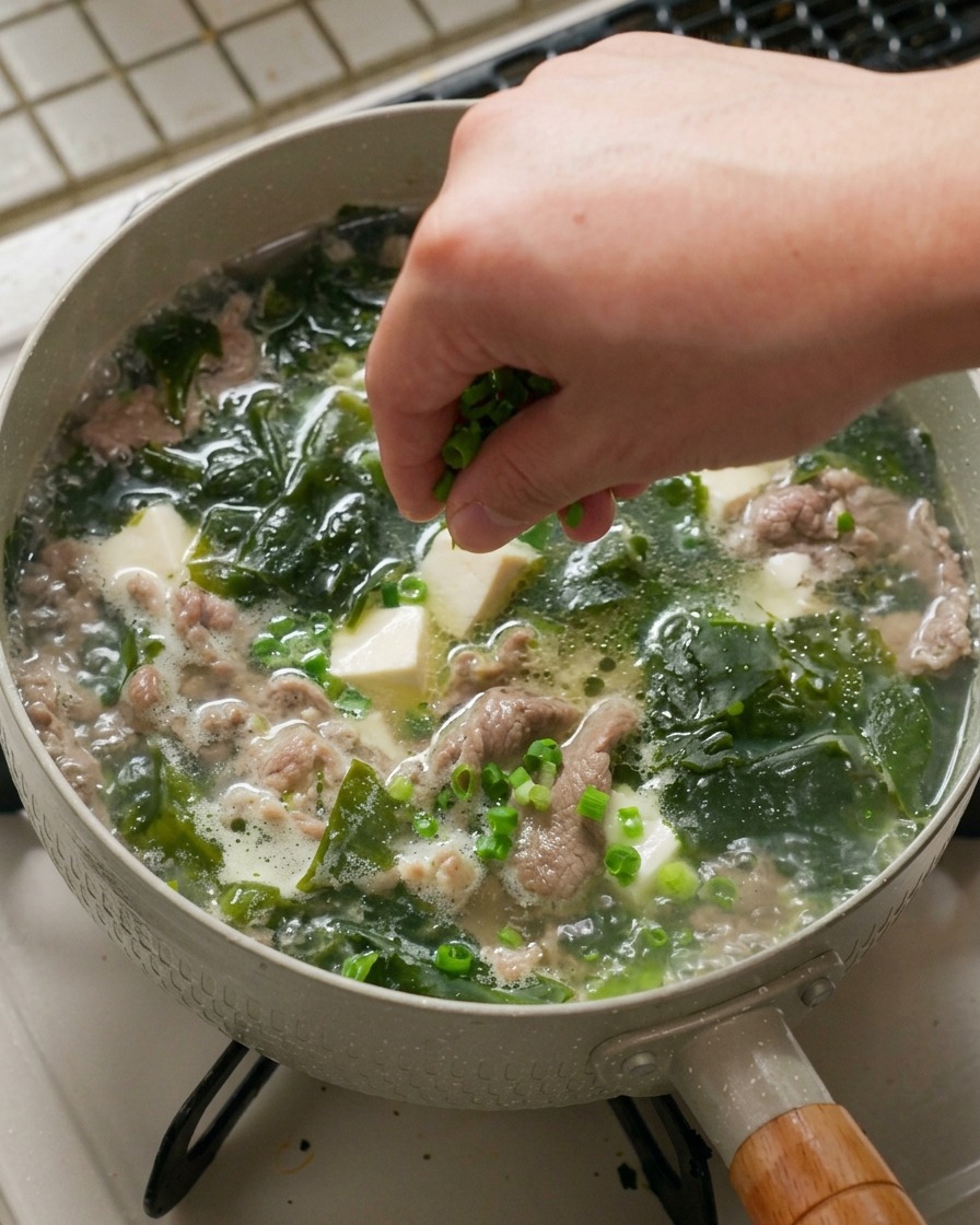 Une main saupoudrant des oignons verts ciselés sur une casserole de soupe chaude.