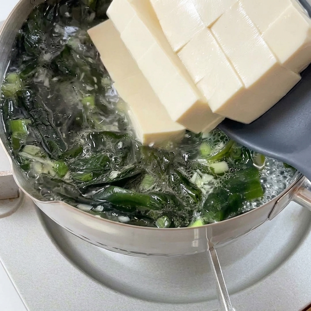 Épaisses tranches de tofu blanc tendre glissant dune spatule grise dans une marmite bouillonnante de bouillon de wakame.