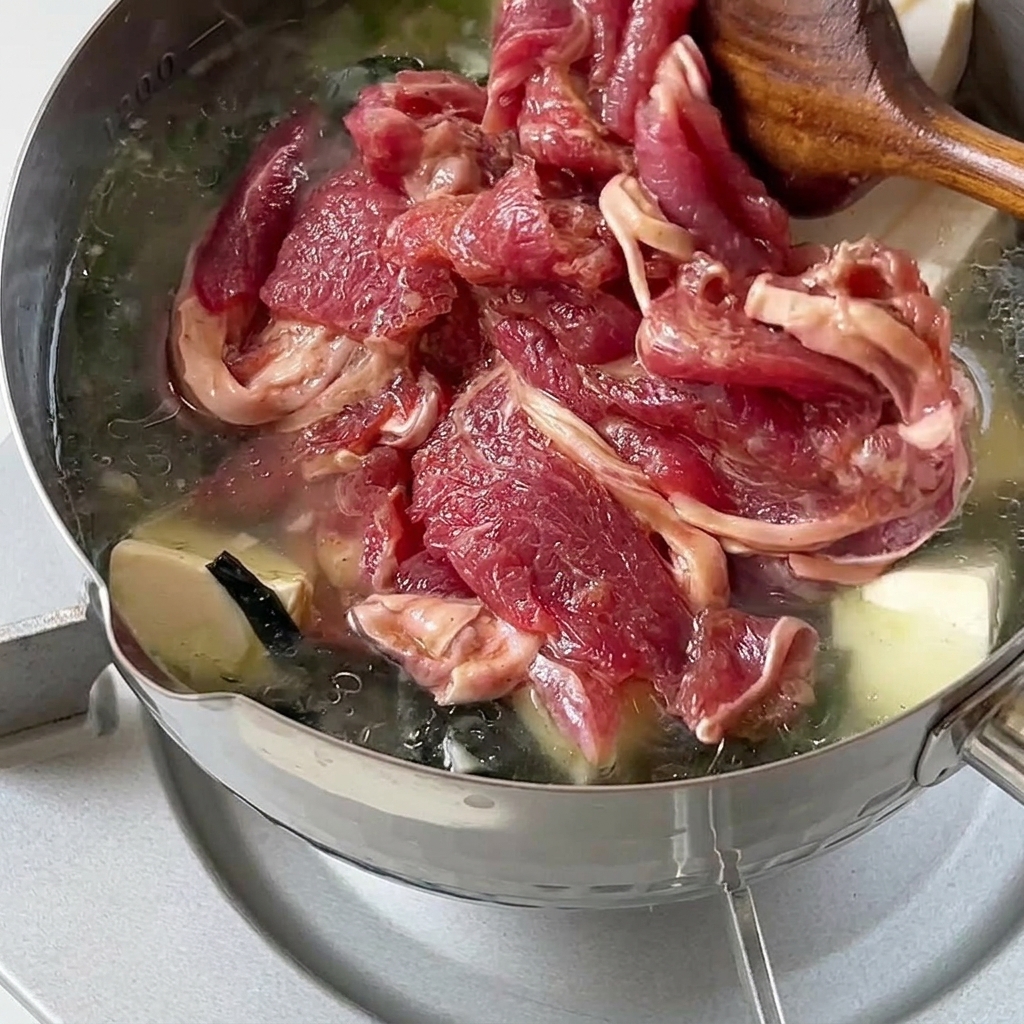 Tranches de bœuf cru mariné ajoutées et remuées délicatement dans une marmite chaude de soupe au wakame et au tofu.