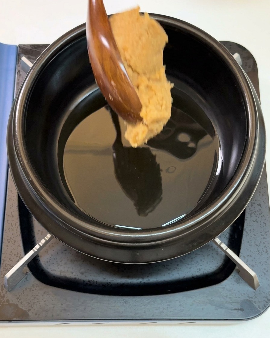 Une grande cuillère en bois versant de lépaisse pâte de soja dans une marmite noire contenant de lhuile de sésame frémissante.