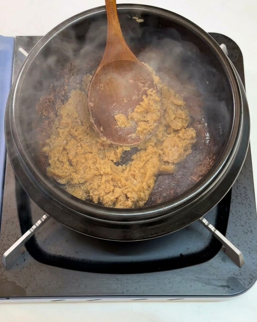 Une cuillère en bois mélangeant de lail haché et de la pâte de soja dans un pot en terre cuite noir sur le feu.