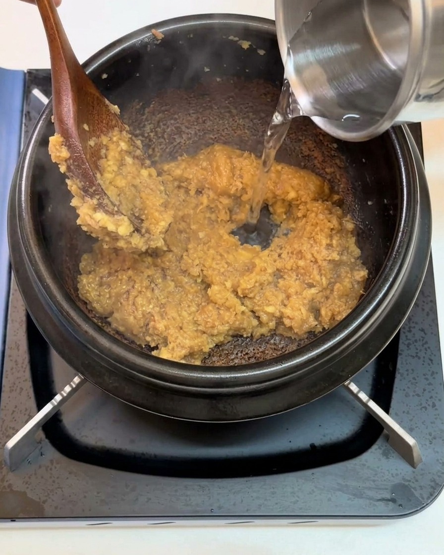 De leau versée depuis un récipient métallique dans une marmite noire contenant la base de pâte de soja.