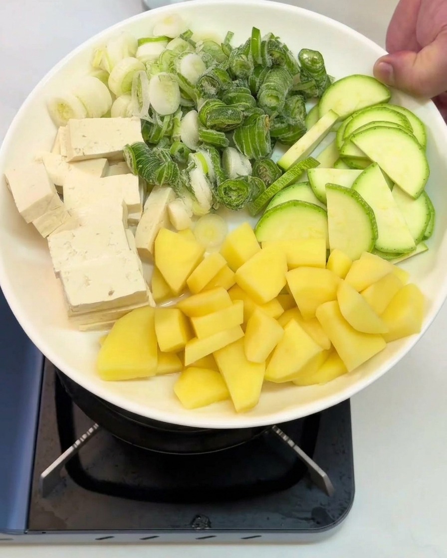 Une assiette blanche tenue au-dessus du feu, présentant des cubes de tofu, des oignons verts émincés, des rondelles de courgette et des dés de pommes de terre.