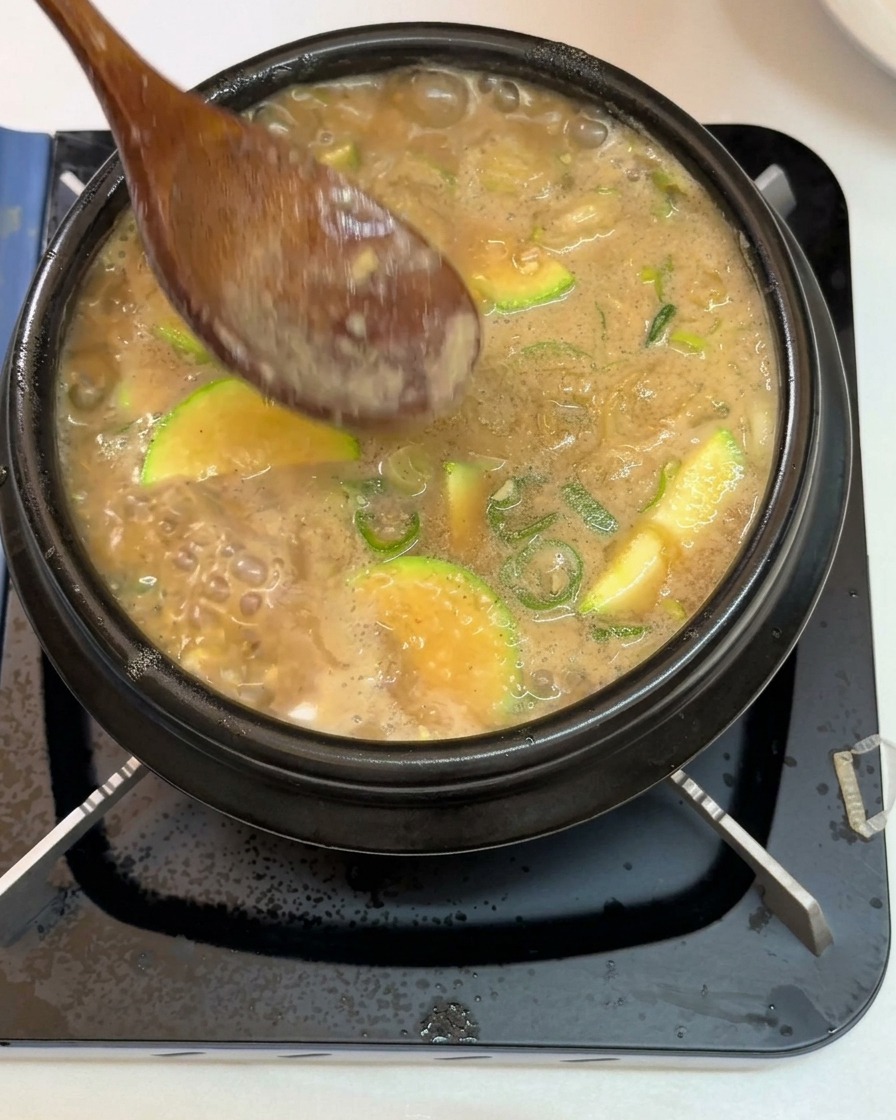 Une cuillère en bois retirant lécume blanche à la surface dun ragoût coréen bouillonnant, garni de courgettes et doignons verts dans un pot en pierre noir.