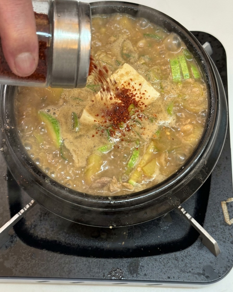 De la poudre de piment rouge saupoudrée depuis un pot en verre et acier inoxydable sur un ragoût coréen frémissant au tofu et aux courgettes.